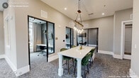 This thoughtfully designed amenity suite occupies a single floor and comprises five distinct areas tailored for both focused work and relaxed collaboration. The space is unified by a consistent design palette of warm neutral tones, patterned carpeting, and clean white millwork that lend it a polished, contemporary feel.

At the heart of the suite is a central lounge and collaborative zone, furnished with a high-top white table and plush emerald green bar chairs beneath a striking industrial-style pendant light fixture with exposed Edison bulbs. Adjacent to this hub, glass-paneled private work pods with black metal framing provide semi-enclosed, quiet workspaces, each outfitted with a desk, task chair, and focused lighting.

A dedicated conference room features a generous light wood table, mesh-back rolling chairs, a wall-mounted display screen, and a textured accent wall. Large windows dressed with plaid drapery invite ample natural light and offer views of the surrounding outdoor greenery.

The lounge seating area provides a more relaxed atmosphere, with leather sofas, accent armchairs, organic wood side tables, and curated artwork. A coffee station makes this corner especially inviting. Floor-to-ceiling windows throughout the suite ensure the space remains bright and connected to the outdoors, creating a warm and productive environment overall.