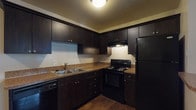 Located at 14417 Hellenic Dr in Tampa, FL, this single-story unit offers a functional and cohesive interior layout. The kitchen is a standout feature, showcasing rich dark espresso cabinetry that extends to the ceiling, granite-style countertops, and a full suite of black appliances including an electric range, built-in dishwasher, and refrigerator. A double-basin stainless steel sink sits beneath ample counter space, making the kitchen both practical and visually striking. Rich, dark hardwood-style flooring runs through the main living areas, providing a warm and consistent aesthetic that flows from the kitchen through the open living space and down the hallway. The living area is generously sized, with neutral-toned walls and a textured ceiling that add depth to the space. A private entrance door opens directly into this area, enhancing accessibility. The bedrooms feature plush carpeting and large windows dressed with vertical blinds, allowing for natural light and outdoor views of the surrounding greenery. The bathroom is clean and well-maintained, featuring a full tub and shower combination with white tiled surroundings, complemented by hardwood-style flooring that ties in with the rest of the home. Overall, the unit presents a neutral, move-in-ready interior with a cohesive design throughout.
