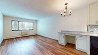 This single-floor residence offers a well-organized layout with distinct living, sleeping, kitchen, and bathroom areas. The open living and dining space is finished with warm, multi-tone hardwood-style flooring that extends throughout much of the unit, reflecting natural light from a large window that frames outdoor views of surrounding trees. An elegant multi-arm chandelier anchors the dining area, which also features a built-in cabinet with granite-style countertop, providing convenient storage and workspace. Adjacent to the entry, the galley-style kitchen is efficiently designed with cream cabinetry, granite-style countertops, a double-basin sink, a dishwasher, electric range, refrigerator, and an over-the-range microwave — all contributing to a functional cooking environment. A louvered closet door near the entry adds practical storage. The bedroom is carpeted for comfort, equipped with a ceiling fan and light fixture, a baseboard heater, and a window overlooking greenery. The full bathroom is finished with white tile, a soaking tub with grab bars and shower, a white vanity with storage, a medicine cabinet mirror, and a classic mosaic tile floor, completing this clean and functional space.