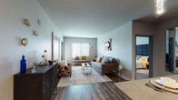 This single-floor residence is thoughtfully designed with a modern aesthetic, blending functionality and style throughout its open-concept layout. Wood-look flooring flows through the main living areas, anchoring the space with warmth and visual continuity.

The kitchen is a standout feature, equipped with stainless steel appliances, dark espresso cabinetry, a tile backsplash, and a peninsula with pendant lighting overhead, offering bar-style seating for casual dining. Recessed lighting and light-toned countertops complete the polished look.

The living room is generously sized and filled with natural light from large windows fitted with horizontal blinds. The open arrangement accommodates multiple seating areas with ample room for a central coffee table and accent furnishings.

The bedroom offers a cozy retreat with plush carpeting, a large upholstered headboard, and a window providing natural light. An additional flex room with carpet flooring and a closet provides versatile living space.

Two bathrooms serve the residence — one featuring a full tub-shower combination with a curved curtain rod and wood-look flooring, and another spacious bathroom accessible via a stylish paneled door. Throughout, neutral wall tones and clean architectural lines create a cohesive, contemporary ambiance.