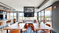Located at 455 Snelling Ave N in Saint Paul, Minnesota, this single-floor amenity space offers a generously sized, thoughtfully designed communal environment for residents. The interior showcases a contemporary aesthetic defined by clean lines, warm wood accents, vertical slatted wood panels, and a refined palette of deep blues and grays complemented by natural light wood flooring.

The living area serves as the social centerpiece, anchored by a striking suspended double-sided glass fireplace framed in bold black metalwork. Floor-to-ceiling windows flood the space with natural light and provide sweeping outdoor views, including a prominent view of a nearby arena. Comfortable seating arrangements, patterned area rugs, and cognac leather chairs create an inviting atmosphere for gathering and relaxation. A foosball table, mounted television, and vending area add recreational variety.

The kitchen is fully equipped with dark granite countertops, warm wood cabinetry, integrated appliances including a wine cooler and microwave, a deep utility sink, and globe pendant lighting — ideal for hosting events. A glass-walled private room offers a quieter retreat or meeting space.

Outside, an expansive terrace features multiple seating zones, a fire pit, pergola with string lights, turf lounging areas, and vibrant outdoor furniture — all set against a striking multi-story building facade of blue metal paneling and masonry.