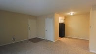 This single-floor unit offers a straightforward, functional layout designed for comfortable everyday living. The main living area is finished with wall-to-wall carpeting in a neutral beige tone, contributing to a warm and understated atmosphere throughout the space. Walls are painted in a soft tan hue, lending a cohesive and clean look to the interior.

The entry area is clearly defined by a solid exterior door equipped with a security lock, with a low-pile entry mat positioned at the threshold. A second interior door, likely leading to a closet or additional room, is situated nearby along the same wall, providing practical storage or utility access.

Adjacent to the living area, the kitchen is partially open to the main space and features dark black appliances, including a full-sized refrigerator, complemented by white upper cabinetry above the counter. The contrast between the dark appliances and light cabinetry creates a functional yet visually defined cooking area. Overhead lighting in the kitchen provides focused illumination, while a flush-mount ceiling fixture serves the living area. Overall, the space presents a clean, move-in ready environment suited for efficient, everyday living.