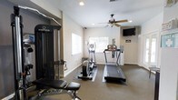 This fitness center offers a clean, functional environment designed to support a well-rounded workout routine. The space is finished with neutral-toned walls and low-pile carpeting, creating a practical and comfortable atmosphere for exercise. Recessed lighting and a ceiling fan with a light fixture provide consistent illumination and airflow throughout the room, ensuring a comfortable environment during workouts.

Natural light enters through multiple windows, including a larger window that offers a view of surrounding greenery, adding a refreshing outdoor connection to the interior space. A wall-mounted flat-screen television is positioned for easy viewing from most areas of the room, making it convenient for entertainment during cardio sessions.

The fitness center is equipped with a treadmill, an elliptical trainer, and a stair climber, all placed on protective floor mats. An Inspire multi-functional cable training system with an adjustable bench anchors the strength training area, providing versatility for a wide range of resistance exercises. A water cooler station is also present, adding a practical amenity for users. The overall layout is thoughtfully organized to maximize usable floor space while keeping equipment accessible and the environment open and uncluttered.