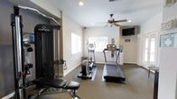 This fitness center offers a clean, functional environment designed to support a well-rounded workout routine. The space is finished with neutral-toned walls and low-pile carpeting, creating a practical and comfortable atmosphere for exercise. Recessed lighting and a ceiling fan with a light fixture provide consistent illumination and airflow throughout the room, ensuring a comfortable environment during workouts.

Natural light enters through multiple windows, including a larger window that offers a view of surrounding greenery, adding a refreshing outdoor connection to the interior space. A wall-mounted flat-screen television is positioned for easy viewing from most areas of the room, making it convenient for entertainment during cardio sessions.

The fitness center is equipped with a treadmill, an elliptical trainer, and a stair climber, all placed on protective floor mats. An Inspire multi-functional cable training system with an adjustable bench anchors the strength training area, providing versatility for a wide range of resistance exercises. A water cooler station is also present, adding a practical amenity for users. The overall layout is thoughtfully organized to maximize usable floor space while keeping equipment accessible and the environment open and uncluttered.
