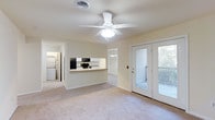 This single-floor residence offers a bright, open layout designed for comfortable everyday living. The main living area features plush wall-to-wall carpeting and a ceiling fan with integrated lighting, creating a welcoming and well-ventilated space. Neutral-toned walls throughout lend a clean, versatile aesthetic that suits a variety of décor styles.

The kitchen is positioned along one wall and features a pass-through counter that connects it to the living space, promoting an open feel while maintaining a functional cooking zone. Black appliances, including a refrigerator and microwave, provide a modern contrast against the light cabinetry. A dedicated laundry area with a stacked washer and dryer unit adds valuable in-unit convenience.

French doors in the living area open to an outdoor deck, allowing natural light to filter into the space and offering a seamless transition between indoor and outdoor living. Views of surrounding greenery are visible through the glass panes, adding a touch of nature to the interior ambiance.

A hallway leads to the additional rooms, with a glimpse of a chandelier-lit space beyond, suggesting a dining area or secondary room. The overall layout is practical and cohesive, making efficient use of the available footprint while maintaining a light, airy atmosphere throughout.