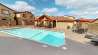 This inviting community space is defined by its warm Spanish-inspired architectural design, characterized by distinctive terracotta tile rooflines, textured stucco exteriors in earthy tones, and wrought iron fencing that frames the outdoor areas with understated elegance. At the heart of the outdoor space sits a generously sized swimming pool with clear, blue-tinted water, complete with lap lane markings and convenient pool entry steps. The surrounding sundeck provides ample room for relaxation, featuring lounge chairs positioned to take in the open sky, as well as multiple seating areas furnished with cast iron tables and chairs shaded by rich, rust-colored patio umbrellas. A dedicated poolside cabana or community building offers additional shelter and amenity access, while storage structures nearby provide functional convenience. A private balcony area, enclosed by sleek vertical metal railings, offers a quiet retreat with views of the community courtyard below. The overall ambiance is sunny, open, and resort-like, with plenty of natural light throughout the day. The cohesive architectural style and well-maintained communal grounds create a relaxed yet polished atmosphere well-suited for outdoor leisure and community gathering.