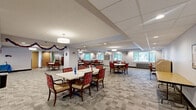 Located at 901 E Como Blvd in Saint Paul, Minnesota, this generously sized single-floor community space offers a welcoming and functional environment designed for social gatherings and shared activities. The open floor plan encompasses a substantial area finished with patterned, neutral-toned carpet tiles that lend a clean, modern aesthetic throughout.

The main gathering area is anchored by multiple round and rectangular tables surrounded by upholstered chairs in rich red and wood-toned finishes, accommodating both small group interactions and larger communal events. A comfortable lounge area features a dark leather sofa and additional seating arranged around a wall-mounted flat-screen television, providing a relaxed setting for informal gatherings.

Natural light flows in through numerous windows fitted with blinds, offering views of mature trees and a well-maintained outdoor green space. Glass-paneled doors provide direct access to the exterior patio and landscaped grounds. A display cabinet, wall clock, and framed artwork contribute warmth and a homey character to the space.

Additional amenities include elevator access via wood-paneled elevator doors, a small reception or service counter, bulletin board areas, and dedicated nooks for quiet activities. Recessed and pendant lighting ensures the space is well-illuminated throughout, creating a bright and inviting atmosphere suitable for a wide range of community programming.