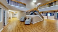 Located at 13910 Hedgewood Dr in Woodbridge, Virginia, this well-appointed community facility is designed to support communal living and shared activities across a single, thoughtfully organized floor. The entrance foyer makes a strong architectural impression with soaring double-height ceilings, warm hardwood-style flooring, and a prominent staircase featuring elegant wood handrails and decorative wrought iron balusters. A cozy seating area with upholstered armchairs is tucked near the base of the stairs, offering a welcoming transition space. The upper-level overlook with matching railing detailing adds a sense of grandeur to the common area below. The expansive dining room accommodates multiple large wooden tables and chairs arranged for group meals or activities, with natural light filtering through large windows dressed in layered curtains. A wall-mounted flat-screen television enhances the space for communal viewing. The fully equipped kitchen is practical and generously sized, featuring extensive warm-toned cabinetry, long stretches of counter space, black appliances including a range and refrigerator, and an open pass-through connecting to adjacent gathering areas. Throughout, the consistent use of light-toned walls, hardwood-style floors, and ample overhead lighting creates a bright, functional, and inviting environment.