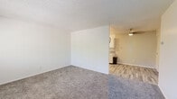 This single-floor unit offers a clean, functional layout designed for comfortable everyday living. The spacious living area is finished with plush grey carpet and crisp white walls, creating a bright and neutral backdrop. Textured ceilings are consistent throughout the carpeted rooms, adding a classic touch to the interior. The living space flows seamlessly toward the kitchen area, where wood-look tile flooring begins, providing a natural visual transition between zones.

The galley-style kitchen is well-appointed with white shaker cabinetry, grey countertops, and a geometric patterned tile backsplash that adds a modern accent. Stainless steel appliances — including a refrigerator, electric range, and dishwasher — are complemented by ample upper and lower cabinet storage and a double basin sink.

The bedroom is generously sized with soft carpet underfoot, a window that brings in natural light and leafy outdoor views, and direct access to a closet. A ceiling fan with pull-chain lighting serves the dining or flex area adjacent to the kitchen.

The full bathroom features a white vanity with a grey countertop, brushed nickel fixtures, a large mirror, a toilet, and a shower-tub combination — all set on wood-look tile flooring that ties the wet areas together cohesively. Overall, the unit presents a move-in ready, well-coordinated living environment.