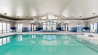 Located at 2025 E Carlson St in Cheyenne, Wyoming, this indoor aquatic facility offers a spacious and well-appointed environment designed for year-round recreational use. The centerpiece of the space is a large, rectangular swimming pool with underwater lighting that illuminates the clear turquoise water, creating a bright and inviting atmosphere. Adjacent to the main pool is a tiled hot tub/spa, accented with blue mosaic tile detailing and accessible via stainless steel handrails. The facility features a vaulted ceiling that rises prominently above the pool area, contributing to an open and airy feel throughout. Exposed HVAC ductwork runs along the ceiling, reflecting the building's functional design aesthetic. A two-toned wall treatment — neutral beige above and slate blue paneling below — adds visual structure to the interior. Large floor-to-ceiling windows line multiple walls, flooding the deck area with natural light and providing views of the surrounding residential community. The generously sized pool deck accommodates multiple seating arrangements, including light-framed chairs and tables, offering comfortable spaces for relaxation. Dedicated entry and exit doors provide convenient access throughout the facility.