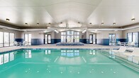 Located at 2025 E Carlson St in Cheyenne, Wyoming, this indoor aquatic facility offers a spacious and well-appointed environment designed for year-round recreational use. The centerpiece of the space is a large, rectangular swimming pool with underwater lighting that illuminates the clear turquoise water, creating a bright and inviting atmosphere. Adjacent to the main pool is a tiled hot tub/spa, accented with blue mosaic tile detailing and accessible via stainless steel handrails. The facility features a vaulted ceiling that rises prominently above the pool area, contributing to an open and airy feel throughout. Exposed HVAC ductwork runs along the ceiling, reflecting the building's functional design aesthetic. A two-toned wall treatment — neutral beige above and slate blue paneling below — adds visual structure to the interior. Large floor-to-ceiling windows line multiple walls, flooding the deck area with natural light and providing views of the surrounding residential community. The generously sized pool deck accommodates multiple seating arrangements, including light-framed chairs and tables, offering comfortable spaces for relaxation. Dedicated entry and exit doors provide convenient access throughout the facility.