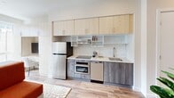 Located at 1314 Spring Garden St in Philadelphia, PA, this modern studio is a single-floor space designed to maximize efficiency without sacrificing style. The open-plan layout seamlessly integrates the living, sleeping, and kitchen areas into one cohesive environment. Clean white walls, recessed lighting, and light hardwood-style flooring create a bright and contemporary ambiance throughout.

The main living area features a large window that draws in abundant natural light and offers views of the surrounding urban streetscape. A built-in storage shelf unit elegantly divides the sleeping area from the living space, while a bold rust-orange sofa adds a warm pop of color against the neutral palette.

The kitchen is well-appointed with light wood upper cabinetry, gray lower cabinets, a white subway tile backsplash, and a full suite of stainless steel appliances including a refrigerator, oven, dishwasher, and flat-top range. Ample upper shelving provides additional storage in this smartly configured culinary space.

The hallway connects the main living area to the private bathroom, which features clean tile flooring, a wall-mounted vanity with backlit mirror, a toilet, and a shower with a crisp white curtain. The overall design is minimal, functional, and polished, making this Spring Garden studio a refined urban retreat.