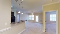 Located at 755 CL Tart Cir in Fayetteville, North Carolina, this single-floor residence offers a well-designed open-concept layout that seamlessly connects the kitchen and living areas. The kitchen features a dark granite-topped breakfast bar illuminated by elegant pendant lighting, providing both a functional workspace and a casual dining option. A refrigerator anchors the kitchen, and neutral-toned cabinetry complements the overall color palette.

The living area is finished with plush carpeting and crowned with crown molding that adds a refined architectural touch throughout. A ceiling fan centered in the living space ensures year-round comfort. Natural light pours in through multiple windows dressed with white blinds, creating a bright and welcoming ambiance. A glass-paneled door leads to the exterior, further enhancing the sense of openness and connectivity to the outdoors.

The walls are painted in soft, neutral tones that contribute to a calm and inviting atmosphere. Additional rooms are accessible via interior doorways, offering private spaces that complement the open communal areas. The consistent use of white trim and molding throughout ties the design together, giving the space a clean and cohesive aesthetic suitable for comfortable everyday living.