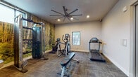 This fitness center offers a comprehensive workout environment designed to accommodate a variety of exercise routines. The space is anchored by a dramatic floor-to-ceiling forest mural that spans one wall, creating an immersive, nature-inspired ambiance that sets it apart from a typical gym setting. A large, multi-blade ceiling fan overhead ensures consistent air circulation throughout the room, complementing the recessed lighting that keeps the space well-lit and inviting.

The cardio zone features a treadmill and an elliptical trainer, both equipped with digital consoles for performance tracking. A stationary bike adds another option for low-impact cardiovascular training. For strength training, the space includes a dual-cable functional trainer, ideal for a wide range of resistance exercises, as well as a dedicated dumbbell rack stocked with a full selection of free weights.

A full-length mirror along one wall expands the sense of space and allows for proper form checks during workouts. The flooring transitions between durable carpet tile in the main exercise area and hard surface flooring near the entryway. Natural light filters in through a window framing an outdoor view of palm trees, reinforcing the connection to the surrounding environment. A water station is also conveniently available within the space.