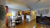 This single-floor home showcases a thoughtfully designed open-concept layout that seamlessly connects the main living areas. The dining area is a welcoming space, highlighted by vaulted ceilings that lend a sense of spaciousness and airiness. A sliding glass door allows natural light to pour in, brightening the room and offering easy access to the outdoors. The dining area features warm hardwood-style flooring that flows consistently throughout the main living spaces, adding a cohesive and inviting feel. A half-wall peninsula connects the dining area to the kitchen, creating an open yet defined transition between the two spaces. The kitchen is well-appointed with warm wood cabinetry, modern appliances including a built-in microwave, and a breakfast bar that provides additional seating and counter space. The textured ceilings add a classic architectural touch, while neutral wall tones keep the space feeling bright and versatile. A hallway off the main area leads to additional rooms, completing this functional and comfortable single-floor layout that balances openness with cozy, lived-in warmth.