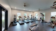 Located at 3085 Highway 123 in San Marcos, Texas, this fitness center offers a dedicated, single-floor exercise room designed to support a well-rounded workout experience. The space features a dark rubberized flooring that is both durable and practical for high-activity use, contrasted against clean white walls that keep the environment bright and open.

Natural light fills the room through a series of large windows along the exterior walls, offering pleasant views of the surrounding greenery and outdoor areas. A set of double glass doors with dark frames provides direct access to an outdoor patio or common area, enhancing ventilation and connectivity to the exterior.

The exercise room is comprehensively equipped with multiple treadmills and an elliptical trainer aligned along the windowed wall for an energizing cardio experience, as well as a stair climber for added variety. Strength training options include a multi-function cable and pulley machine, a weight bench, and a plate-loaded leg press or rowing machine. Wall-mounted mirrors expand the sense of space and allow users to monitor their form. Recessed ceiling lighting and a decorative ceiling fan complete the functional yet polished aesthetic, making this fitness center a practical and inviting amenity.