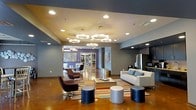 This single-floor space presents an open, contemporary layout with a strong emphasis on modern design and functionality. The main lounge area is generously proportioned, featuring polished concrete flooring that adds an industrial yet refined aesthetic throughout. A striking arrangement of circular pendant chandeliers serves as a focal point overhead, complemented by recessed lighting that ensures the space is well-illuminated and inviting.

The seating area is thoughtfully arranged with a clean-lined white sofa accented with decorative pillows, paired with sleek swivel chairs in a deep plum tone. Cylindrical ottomans in metallic silver and navy tones are scattered across a vibrant multicolored area rug, adding both practicality and a playful design element. The walls are painted in a cool, muted gray-blue palette, grounding the space with a calm, cohesive tone.

In the background, a secondary seating area is visible beyond an open passageway, suggesting multiple functional zones within the layout. A kitchen or kitchenette area is partially visible on the right, equipped with stainless steel appliances. Decorative wall art and maps add character and visual interest throughout the space.