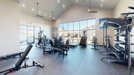 Located at 2751 Ruby River Drive in Lexington, Kentucky, this single-floor fitness center offers a comprehensive workout facility designed with both function and comfort in mind. The main exercise room is an impressive, open space distinguished by soaring vaulted ceilings, recessed lighting, and multiple ceiling fans that keep the environment well-ventilated. Expansive floor-to-ceiling windows line one wall, flooding the room with natural light and providing views of the surrounding neighborhood. The durable dark rubber flooring underfoot is well-suited for heavy equipment and high-intensity training. The facility is outfitted with a robust selection of cardio machines, including treadmills, ellipticals, and stair climbers, alongside a comprehensive strength training area featuring cable machines, weight benches, a multi-functional cable tower, and organized dumbbell racks with medicine balls. Wall-mounted televisions are positioned for convenient viewing during workouts. A secondary exercise area provides additional workout space, while a transitional hallway connects to a warmly lit sauna room, accented by natural wood paneling. A dual-basin water fountain station is conveniently located near the entrance, and large glass exterior windows allow natural light to carry through the space. Together, these amenities create a fully equipped, inviting fitness environment.