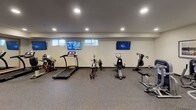 Located at 568 Settlers Ridge Pkwy in Woodbury, Minnesota, this fitness center is a thoughtfully designed, single-floor facility built to accommodate a wide range of exercise needs. The space features durable speckled rubber flooring throughout, providing both safety and comfort during workouts.

The cardio zone is lined with multiple machines, including treadmills, stationary bikes, and a rowing machine, positioned along a wall with small windows that allow natural light to filter in. Mounted flat-screen televisions are installed above the cardio equipment, offering entertainment during exercise sessions.

The strength training area includes a selection of Life Fitness selectorized machines, such as a squat machine and additional cable-based units, enabling targeted muscle training. A dedicated free weights section features a rack stocked with dumbbells, medicine balls, and an adjustable weight bench, offering versatility for functional training.

A large wall-to-wall mirror spans one section of the room, enhancing the sense of space and supporting proper form during workouts. Recessed lighting throughout ensures the space is well-illuminated. The muted blue and neutral wall palette contributes to a focused, energizing atmosphere. An adjacent lounge area is visible through a glass-paneled door.