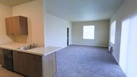 This single-floor space offers an open-concept design that integrates the kitchen and living areas into one cohesive, functional layout. The kitchen is positioned along a partial wall partition, featuring a light-toned countertop, an undermount sink, and wood-finish cabinetry below, providing practical storage. The countertop extends slightly, creating a natural transition between the kitchen and the expansive living area beyond.

The living space is generously sized, finished with plush wall-to-wall carpeting in a neutral gray tone that adds warmth and comfort underfoot. Crisp white and light beige walls keep the interior bright and versatile, complementing a range of décor styles. A window on the far wall allows natural light to fill the room, enhancing the open and airy feel of the space.

Tall ceilings in the kitchen zone contribute to a sense of spaciousness, while the open floor plan allows for flexible furniture arrangements. A doorway visible in the background suggests access to additional areas of the unit. Overall, the space presents a clean, modern aesthetic with a neutral palette well-suited for a variety of lifestyles.
