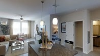This single-story space showcases an open-concept design that connects the kitchen, living, and bedroom areas in a cohesive, functional layout. The kitchen island serves as a central focal point, featuring dark granite countertops and two elegant pendant lights that hang at varying heights, casting a warm, inviting glow. Cabinetry beneath the island provides ample storage, while the overall kitchen design emphasizes both style and practicality.

The open layout allows for a clear sightline into the living area, where a ceiling fan contributes to comfortable airflow. Natural light filters in through a window dressed with neutral curtains, brightening the space and enhancing its airy feel. The flooring transitions smoothly throughout, reinforcing the connected nature of the floor plan.

A bedroom is visible from the kitchen area, accessible through a doorway framed by crisp white trim. The bedroom features warm ambient lighting and is accented by framed wall art nearby. An additional doorway hints at further utility spaces, such as a closet or hallway. The neutral color palette throughout creates a calm, cohesive atmosphere well-suited for everyday living.