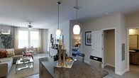 This single-story space showcases an open-concept design that connects the kitchen, living, and bedroom areas in a cohesive, functional layout. The kitchen island serves as a central focal point, featuring dark granite countertops and two elegant pendant lights that hang at varying heights, casting a warm, inviting glow. Cabinetry beneath the island provides ample storage, while the overall kitchen design emphasizes both style and practicality.

The open layout allows for a clear sightline into the living area, where a ceiling fan contributes to comfortable airflow. Natural light filters in through a window dressed with neutral curtains, brightening the space and enhancing its airy feel. The flooring transitions smoothly throughout, reinforcing the connected nature of the floor plan.

A bedroom is visible from the kitchen area, accessible through a doorway framed by crisp white trim. The bedroom features warm ambient lighting and is accented by framed wall art nearby. An additional doorway hints at further utility spaces, such as a closet or hallway. The neutral color palette throughout creates a calm, cohesive atmosphere well-suited for everyday living.