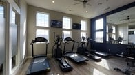 Located at 7505 Worthington Galena Rd in Columbus, Ohio, this single-floor fitness center is designed to provide a functional and motivating environment for residents and guests alike. The space is outfitted with a selection of cardio equipment, including treadmills, elliptical trainers, and a stair climber, all arranged to maximize floor space and allow comfortable circulation between machines. The room features light wood-style flooring that adds a clean, modern aesthetic underfoot. Large, multi-pane windows line the walls on multiple sides, flooding the interior with generous natural light and offering open views of the surrounding area — creating an airy, welcoming atmosphere. A ceiling fan overhead ensures steady air circulation during workouts, while recessed lighting supplements the natural light for consistent illumination throughout the day. Wall-mounted flat-screen televisions are positioned for easy viewing from the cardio machines, enhancing the workout experience. An accent wall in deep navy blue adds a bold design element, contrasting with the neutral tones of the remaining walls and contributing to the space's contemporary feel. Overall, this fitness center combines practical equipment with thoughtful design details to create a comfortable and well-lit exercise environment.