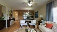 This single-story space showcases a thoughtfully designed open living area that blends comfort with contemporary style. The main living space features warm hardwood-style flooring that extends throughout, anchoring the room with a natural, cohesive aesthetic. A ceiling fan with a built-in light fixture is centrally positioned, providing both air circulation and ambient illumination.

The living area is furnished with a pair of patterned accent chairs, a cowhide-style area rug, and a sofa, creating a cozy yet sophisticated seating arrangement. A large mirrored wall panel serves as a striking focal point on the accent wall, which is painted in a warm taupe tone that adds depth and contrast to the lighter surrounding walls. Decorative plants and artwork further enhance the space's visual appeal.

A pendant light fixture marks a defined transition into the adjacent dining or secondary area, while a console table near the entryway provides additional functionality. The entryway, visible through the open layout, features a classic arched transom window above the front door, allowing natural light to filter into the space.