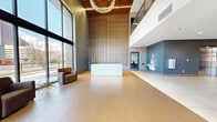 This contemporary lobby hallway presents an architecturally striking first impression through its double-height ceilings and expansive floor-to-ceiling glazing that frames panoramic views of the surrounding urban skyline. Warm-toned large-format ceramic tiles extend across the open floor, anchoring the generous, airy footprint of the space.

A dramatic vertical wood slat accent wall serves as the lobby's central design focal point, rising the full height of the interior alongside a sleek white reception desk positioned at its base. An elegant chandelier descends from a warm wood-paneled ceiling detail above, adding a refined, decorative element to the otherwise clean-lined aesthetic.

A dedicated seating lounge adjacent to the glazed facade features upholstered armchairs in yellow and brown tones arranged around a gold starburst side table, offering a welcoming area for visitors. The hallway connects to a secondary corridor housing stainless steel elevator banks framed by cool grey walls and light-toned tiles.

Exposed brick near the building's entrance doors adds textural warmth, complementing framed artwork displayed along the walls. A water fountain station and additional utility fixtures are thoughtfully integrated into the layout. Overall, the lobby conveys a polished, professional, and welcoming ambiance with a well-balanced mix of modern and warm materials.