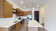 This single-floor unit showcases a contemporary design aesthetic with clean lines and a neutral palette throughout. The kitchen serves as a central highlight, featuring warm wood-toned cabinetry with black hardware that contrasts elegantly against crisp white quartz countertops. A full suite of stainless steel appliances includes a French door refrigerator with ice dispenser, an electric range with a stainless steel chimney hood, a built-in microwave, and a dishwasher. A spacious central island with a white countertop provides additional prep and dining space, while glass-front upper cabinets and an integrated wine rack add functional character. Pendant lighting over the island and recessed ceiling lights illuminate the space, which is grounded by durable wood-look flooring. The bathroom presents a clean, minimal design with white flat-panel doors featuring black hardware, wood-toned cabinetry with a white countertop, and a tiled tub-shower combination with a subway tile surround. Wood-look flooring carries through into this space as well. The bedroom is finished with soft grey carpet, bright white walls, and a large window that allows generous natural light and views of the surrounding urban streetscape.