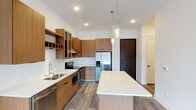 This single-floor unit showcases a contemporary design aesthetic with clean lines and a neutral palette throughout. The kitchen serves as a central highlight, featuring warm wood-toned cabinetry with black hardware that contrasts elegantly against crisp white quartz countertops. A full suite of stainless steel appliances includes a French door refrigerator with ice dispenser, an electric range with a stainless steel chimney hood, a built-in microwave, and a dishwasher. A spacious central island with a white countertop provides additional prep and dining space, while glass-front upper cabinets and an integrated wine rack add functional character. Pendant lighting over the island and recessed ceiling lights illuminate the space, which is grounded by durable wood-look flooring. The bathroom presents a clean, minimal design with white flat-panel doors featuring black hardware, wood-toned cabinetry with a white countertop, and a tiled tub-shower combination with a subway tile surround. Wood-look flooring carries through into this space as well. The bedroom is finished with soft grey carpet, bright white walls, and a large window that allows generous natural light and views of the surrounding urban streetscape.