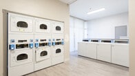 Located at 6303 Indian School Rd NE in Albuquerque, New Mexico, this single-floor shared laundry facility is designed for practical, efficient use. The space is finished with light wood-look luxury vinyl flooring throughout, lending a clean and modern aesthetic that is easy to maintain.

The main laundry area houses a row of front-loading top-control washing machines lined along one wall, illuminated by natural light streaming through floor-to-ceiling sheer-curtained windows. Opposite the washers, a bank of stacked commercial dryers—numbered for easy identification and equipped with digital payment interfaces—provides ample drying capacity for multiple users simultaneously. A rolling laundry cart is available for convenience.

A dedicated waiting and folding area adjoins the laundry machines, furnished with red upholstered armchairs and a long folding table, offering a comfortable space to sit while waiting or to sort and fold laundry. Informational signage, including app-based payment instructions, is posted on the walls for user guidance.

The facility is brightly lit with recessed and overhead fluorescent lighting, complementing the natural light from the large windows. Neutral gray-white walls and clean lines throughout contribute to an organized, welcoming environment suited for communal use.