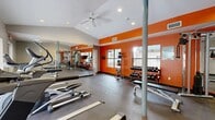 This single-level fitness center is designed to provide a comprehensive workout environment with a energetic, motivating aesthetic. Bold orange accent walls contrast against neutral gray tones, creating a visually stimulating atmosphere throughout the space. The ceiling features recessed lighting and a ceiling fan, ensuring the room remains comfortable and well-lit during workouts.

The cardio zone is anchored by a large floor-to-ceiling mirror wall that visually expands the space and includes treadmills and an elliptical trainer, positioned to overlook a large window with direct views of an outdoor pool area, adding a refreshing outdoor connection. The strength training area is well-stocked with a tiered dumbbell rack offering a full range of free weights, a medicine ball storage rack, a multi-station cable machine, and a workout bench. The durable gray flooring throughout provides a practical, low-maintenance surface suitable for high-traffic use.

Natural light enters through multiple windows along the orange accent wall, complementing the recessed overhead lighting. The layout is open and spacious, offering ample room to move freely between equipment zones, making this fitness center a functional and inviting space for a variety of workout routines.