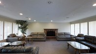 This single-floor space centers around a generously sized living area designed for comfort and easy gathering. The room's most notable feature is a classic brick fireplace positioned along the main wall, serving as a natural focal point and adding warmth and character to the space. Flanking the fireplace are two large, plush gray leather sofas, complemented by additional seating and accent tables that create an inviting, well-furnished arrangement. The room is finished with wall-to-wall neutral carpeting that contributes to a warm and cozy atmosphere. Recessed lighting is distributed evenly across the ceiling, providing consistent illumination throughout the space, while natural light filters in through a window on the side wall. Crown molding along the ceiling adds a subtle architectural detail that elevates the overall finish of the room. A large indoor tree in the corner introduces a touch of greenery, softening the space and adding visual interest. Overall, the living area strikes a balance between classic design elements and functional comfort, making it a welcoming environment suited for both relaxed everyday use and social gatherings.