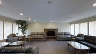 This single-floor space centers around a generously sized living area designed for comfort and easy gathering. The room's most notable feature is a classic brick fireplace positioned along the main wall, serving as a natural focal point and adding warmth and character to the space. Flanking the fireplace are two large, plush gray leather sofas, complemented by additional seating and accent tables that create an inviting, well-furnished arrangement. The room is finished with wall-to-wall neutral carpeting that contributes to a warm and cozy atmosphere. Recessed lighting is distributed evenly across the ceiling, providing consistent illumination throughout the space, while natural light filters in through a window on the side wall. Crown molding along the ceiling adds a subtle architectural detail that elevates the overall finish of the room. A large indoor tree in the corner introduces a touch of greenery, softening the space and adding visual interest. Overall, the living area strikes a balance between classic design elements and functional comfort, making it a welcoming environment suited for both relaxed everyday use and social gatherings.