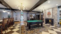 Located at 891 Ingleside Ave in Columbus, Ohio, this single-floor amenity space offers a sophisticated and eclectic atmosphere designed for socializing, entertainment, and productivity. The game room serves as a central focal point, showcasing a full-size billiards table set atop a bold black-and-white checkered tile floor, complemented by a bar-height counter with classic wooden stools and striking geometric gold chandeliers overhead. Dark exposed beams run throughout, adding architectural definition and contrast against light walls and warm wood flooring found in adjacent areas. A spacious lounge area features an eclectic mix of velvet and upholstered seating in rich jewel tones, arranged beneath a coffered ceiling with large floor-to-ceiling windows that flood the space with natural light and offer outdoor views. A dedicated screening room provides an intimate cinematic experience with deep navy sectionals, colorful accent pillows, warm sconce lighting, teal-paneled walls, and a large projection screen. The catering kitchen corridor is outfitted with dark cabinetry, white countertops, and direct access to an outdoor pool terrace. A business center rounds out the offerings, featuring workstations, pendant lighting, and a comfortable seating nook, all framed by textured brick accent walls throughout the space.