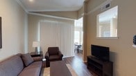 This single-floor space showcases an open-concept layout that connects the living area to the dining space, creating a cohesive and functional flow. The living room is appointed with dark-upholstered seating, including a plush armchair and sofa, centered around a wooden coffee table atop a textured area rug. Hardwood-style flooring extends throughout, adding warmth and continuity to the space.

Large vertical blinds cover a full-height sliding glass door, allowing for adjustable natural light while offering access to the exterior. Crown molding and a subtle tray ceiling detail add architectural character to the living area, while warm beige walls contribute to a neutral, inviting ambiance.

A structural column with decorative molding separates the living and dining zones, maintaining an open feel while subtly defining each space. The dining area, visible through the opening, features a dark table with upholstered chairs and benefits from additional natural light via a nearby window. Track lighting overhead provides supplemental illumination. A flat-screen television is positioned on a dark media console along one wall, completing the functional and well-organized living space.