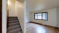 This three-floor space presents a straightforward and functional design with clean, neutral interiors throughout. The main living area visible in the image features light-toned hardwood flooring that adds warmth and continuity to the space, complemented by crisp white walls and standard-height ceilings. A large window with dark framing allows natural light to filter into the room, offering views of the surrounding exterior landscape including neighboring structures and mature trees. Vertical blinds provide privacy control at the window.

A carpeted staircase with a structural dividing wall separates the living area from the stairwell, creating a defined transition between levels. The staircase leads to additional floors above, extending the usable space across three levels in total. A floor-level ventilation register is visible along the baseboard, indicating a forced-air heating and cooling system. The overall aesthetic is minimal and move-in ready, with neutral tones throughout that provide a versatile backdrop adaptable to various interior styles. The multi-level layout offers distinct separation of living spaces across the three floors.