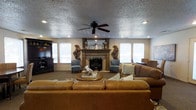 This single-floor residence centers around a generously sized living area designed for both comfort and style. The room's focal point is a classically styled wood-mantel fireplace, flanked by a pair of elegant accent chairs and decorative wall-mounted log arrangements that add a rustic, natural touch. A large tan leather sofa faces the fireplace, anchored by a round wooden coffee table that ties the seating arrangement together.

Recessed lighting throughout the ceiling is complemented by a ceiling fan with a dark finish, ensuring the space remains well-lit and comfortable year-round. The textured ceiling adds depth and character to the room. Multiple windows along the walls flood the interior with natural light, creating a bright and inviting atmosphere.

To one side, a dark wood media console provides dedicated entertainment space, while a glimpse of a dining area is visible in the background, suggesting a well-connected, open layout. The neutral-toned carpeted flooring and warm wall colors contribute to the overall cohesive and welcoming ambiance. Thoughtful decor details, including decorative trays, floral accents, and patterned throw pillows, round out the space with a curated, lived-in charm.