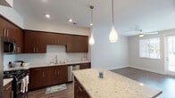 This single-floor residence showcases a thoughtfully designed open-concept layout connecting the kitchen, dining, and living areas. The kitchen is a standout feature, equipped with rich dark-walnut cabinetry, light granite countertops, and a generous central island illuminated by elegant teardrop pendant lights. Stainless steel appliances — including a gas range, built-in microwave, French door refrigerator, and dishwasher — provide a fully functional cooking space. Wood-look plank flooring runs throughout the main living areas, adding warmth and continuity. A stacked washer and dryer unit is conveniently integrated near the kitchen. The living area benefits from natural light through large windows and a glass-paneled exterior door, complemented by a ceiling fan overhead. The bedroom offers plush carpeting, dual windows with blinds, and its own ceiling fan, creating a comfortable, well-lit retreat. The bathroom features a combined tub and shower with a curved rod, built-in shelving, wood-look flooring, and clean neutral finishes. Throughout the unit, neutral wall tones and recessed lighting contribute to a bright, cohesive, and contemporary ambiance.