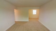 This single-floor residential space offers a clean and straightforward layout designed for comfortable everyday living. The interior is finished with soft, neutral wall tones in warm cream and beige hues, creating a bright and welcoming atmosphere throughout. Wall-to-wall carpeting in a sandy tan shade runs across the floors, providing a cozy and uniform feel underfoot.

The living area flows openly into an adjacent connected room, separated by a partial wall that defines each space while maintaining an open, airy quality. The layout allows for flexible furniture arrangements and easy movement between areas. A window in the adjoining room invites natural light into the space, contributing to its warm, livable ambiance.

Clean white baseboards line the perimeter of each room, adding a crisp contrast to the neutral walls and carpeting. The ceilings are smooth and finished in white, complementing the overall light color palette. Light switches and electrical fixtures are thoughtfully positioned throughout, reflecting a practical and functional design.

Overall, this space presents a well-maintained, move-in-ready interior with a simple, consistent aesthetic. Its open connectivity between rooms, neutral finishes, and natural light make it a versatile and comfortable place to call home.