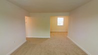 This single-floor residential space offers a clean and straightforward layout designed for comfortable everyday living. The interior is finished with soft, neutral wall tones in warm cream and beige hues, creating a bright and welcoming atmosphere throughout. Wall-to-wall carpeting in a sandy tan shade runs across the floors, providing a cozy and uniform feel underfoot.

The living area flows openly into an adjacent connected room, separated by a partial wall that defines each space while maintaining an open, airy quality. The layout allows for flexible furniture arrangements and easy movement between areas. A window in the adjoining room invites natural light into the space, contributing to its warm, livable ambiance.

Clean white baseboards line the perimeter of each room, adding a crisp contrast to the neutral walls and carpeting. The ceilings are smooth and finished in white, complementing the overall light color palette. Light switches and electrical fixtures are thoughtfully positioned throughout, reflecting a practical and functional design.

Overall, this space presents a well-maintained, move-in-ready interior with a simple, consistent aesthetic. Its open connectivity between rooms, neutral finishes, and natural light make it a versatile and comfortable place to call home.