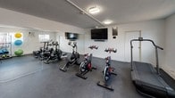 This fitness center is a comprehensively outfitted single-level facility designed to support a wide range of workout routines. The space features white walls, a textured ceiling, and durable dark rubber flooring throughout, creating a clean and functional environment suited for regular physical activity.

The cardio area is equipped with multiple spin bikes and at least one treadmill, positioned to face wall-mounted flat-screen televisions that provide entertainment during workouts. A large wall mirror along one side of the room expands the sense of space while allowing users to monitor their form. Flush-mounted ceiling lights ensure consistent, even illumination throughout.

A dedicated functional training zone houses an organized rack stocked with stability balls in various sizes, resistance bands, and exercise mats. TRX suspension straps are mounted to a wall-anchored rig, and a shelving unit holds kettlebells and other accessories. Additional equipment includes a battle rope and balance tools, offering versatility for strength and conditioning work.

Natural light enters through windows on the exterior walls, supplementing the artificial lighting. The overall layout is efficient and well-organized, making effective use of the available space to accommodate multiple users and training modalities simultaneously.