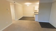 This single-floor unit offers a functional and straightforward layout designed to maximize usability throughout its open living spaces. The main living and dining area is finished with plush carpeting in a neutral tone, creating a warm and comfortable atmosphere. A transition to dark flooring marks the dining zone, providing a subtle visual distinction between the two areas. Soft, cream-colored walls are accented by white chair rail molding that runs along the perimeter, adding a classic architectural detail to the interior. Ceiling-mounted lighting provides consistent illumination throughout the space. The kitchen is conveniently positioned adjacent to the dining area and features white upper cabinetry paired with dark countertops, offering a clean and practical workspace. A pass-through or peninsular counter connects the kitchen to the adjoining area, promoting an open flow between spaces. White baseboards and trim throughout the unit provide a crisp, finished look. Ceiling vents indicate a central HVAC system for year-round climate control. Overall, the space presents a clean, move-in ready environment with a neutral palette well-suited for a variety of interior styles and furnishings.