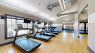 Located at 6400 Preserve Crossing Blvd N in Gahanna, Ohio, this generously sized fitness center offers a comprehensive, multi-zone exercise facility designed to accommodate a wide range of workout needs. The space is organized into distinct functional areas connected by a clean, well-lit hallway finished with warm hardwood-style flooring and neutral gray walls.

The cardio zone features a long row of treadmills positioned alongside large windows that frame views of the surrounding landscape, filling the area with natural light. Durable rubber flooring throughout the workout areas ensures safety and comfort during high-impact activities. A dedicated cycling and elliptical section provides additional low-impact cardio options, also benefiting from multiple windows overlooking the exterior.

The strength training area is well-equipped with a full rack of dumbbells, weight benches, cable machines, and selectorized strength equipment, all arranged to maximize floor space and usability. Large mirrors line the walls, enhancing the sense of openness.

A separate studio space features vaulted ceilings with exposed ductwork, a ballet barre along one wall, and open floor space suitable for group fitness, stretching, or functional training. Storage for mats and fitness balls is built into the studio. Overhead linear lighting throughout ensures the entire facility is bright and inviting, creating a motivating environment for all levels of fitness.