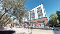 This expansive urban plaza is set within a lively mixed-use development, characterized by a striking blend of red brick and white-paneled multi-story buildings that frame the open-air space. The ground level of the surrounding structures features large storefront windows with decorative awnings, complemented by an outdoor patio area furnished with bistro-style tables, chairs, and retractable red umbrellas — ideal for al fresco dining and socializing.

The plaza itself is paved with broad, light-toned concrete tiles that extend generously in multiple directions, creating a sense of openness and connectivity. Classic-style lamp posts punctuate the walkways, adding a timeless aesthetic to the urban environment. Mature olive trees, tall bamboo hedges, and large sculptural planters filled with succulents and mixed greenery provide natural contrast against the hard architectural surfaces, offering shade and a sense of tranquility.

Notable neighboring elements include a restaurant identified as Muzeo, visible signage for Capitol Plaza, and a Courtyard hotel, suggesting the plaza serves as a central hub within a mixed retail, dining, and hospitality district. The surrounding cityscape, with palm trees and modern high-rises visible in the background, reinforces the plaza's urban character. Under clear blue skies, the space is bright, welcoming, and well-suited for pedestrian activity and community engagement.