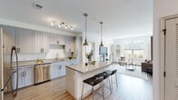 This well-appointed single-floor residence offers a thoughtfully designed open-concept layout that seamlessly connects the kitchen, dining, and living areas. The kitchen is a standout feature, showcasing gray shaker-style cabinetry, granite countertops, and a full suite of stainless steel appliances including a refrigerator, range, microwave, and dishwasher. A central island with seating provides additional prep space and serves as a natural gathering point, accented by stylish pendant lighting overhead. Light wood-style flooring flows throughout the main living areas, enhancing the bright and airy atmosphere. The living room is generously proportioned, featuring large windows that invite abundant natural light and provide views of the outdoors, along with access to an exterior door. A hallway connects the main living space to the private quarters, which include two carpeted bedrooms with clean white walls and ample closet storage. The bathrooms are finished with large-format marble-style tile flooring, gray vanity cabinetry, and modern fixtures, contributing to the overall contemporary aesthetic. Throughout the unit, a neutral color palette and modern hardware choices create a cohesive and polished look that is both functional and visually appealing.