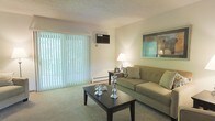 This multi-floor residential space offers a well-organized layout spread across three levels, providing generous room to live and move comfortably. The living area serves as the heart of the home, featuring soft wall-to-wall carpeting that adds warmth underfoot and a cozy ambiance. A large sliding glass door dressed with vertical blinds floods the room with natural light and offers a bright, airy feel. A wall-mounted air conditioning unit and a baseboard heating system ensure year-round climate comfort. The neutral, light-toned walls create a clean and versatile backdrop, complemented by a textured ceiling that adds subtle character to the space. The living room accommodates a classic seating arrangement, with room for a sofa, accent pillows, a coffee table, and a side table with a lamp, making it an inviting area for relaxation or entertaining. The overall aesthetic is understated and functional, with a palette of soft greens and warm neutrals that lend a calm, welcoming atmosphere throughout. With three floors to explore, this space offers ample opportunity for dedicated living, sleeping, and utility areas, making it a practical and comfortable place to call home.
