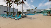 This expansive outdoor recreational area boasts a resort-like atmosphere with a variety of amenities. The centerpiece is a large, curved swimming pool with crystal-clear blue water, surrounded by a spacious concrete deck. Adjacent to the pool is a unique water play area featuring a pirate ship structure and spray fountains, perfect for children's entertainment. 

The space is dotted with numerous turquoise lounge chairs, providing ample seating for sunbathing and relaxation. Tall palm trees line the area, offering natural shade and a tropical ambiance. Other vegetation includes various shrubs and small trees, creating a lush, green environment.

The facility includes a large, white metal building with windows, likely housing indoor amenities or services. A wooden fence partially encloses the area, offering privacy while still allowing views of the surrounding landscape. Lighting fixtures are strategically placed throughout, ensuring the space can be enjoyed into the evening hours.

Additional features include umbrellas for shade, a central planter with decorative elements, and a mix of concrete and textured surfaces for visual interest and functionality. The overall design creates a harmonious blend of recreational facilities and natural elements, providing a versatile space for leisure and enjoyment.