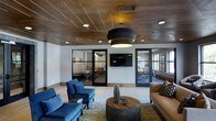 Located in Dallas, this single-story space showcases a thoughtfully curated interior that balances modern aesthetics with warm, industrial-chic character. The main lounge area immediately draws attention with its striking rustic wood-plank ceiling, accented by recessed lighting and a bold, tiered black drum pendant light that serves as a focal centerpiece. Light-toned hardwood flooring contrasts beautifully with the rich ceiling overhead, creating a layered visual depth throughout the space.

Seating in the lounge is arranged around a round, mixed-material coffee table, featuring deep navy blue accent chairs and a tan leather sofa with patterned throw pillows—offering both comfort and stylistic contrast. A wall-mounted flat-screen television anchors one wall, adding a functional element to the social area.

Glass-paneled doors with black steel frames flank the lounge on both sides, providing visibility into adjacent rooms that appear to function as private meeting or conference spaces. These rooms are furnished with office-style seating and tables, allowing for flexible use. The transparent partitions maintain an open, connected feel while defining distinct functional zones. Overall, the space exudes a professional yet inviting ambiance suited for collaboration and gathering.